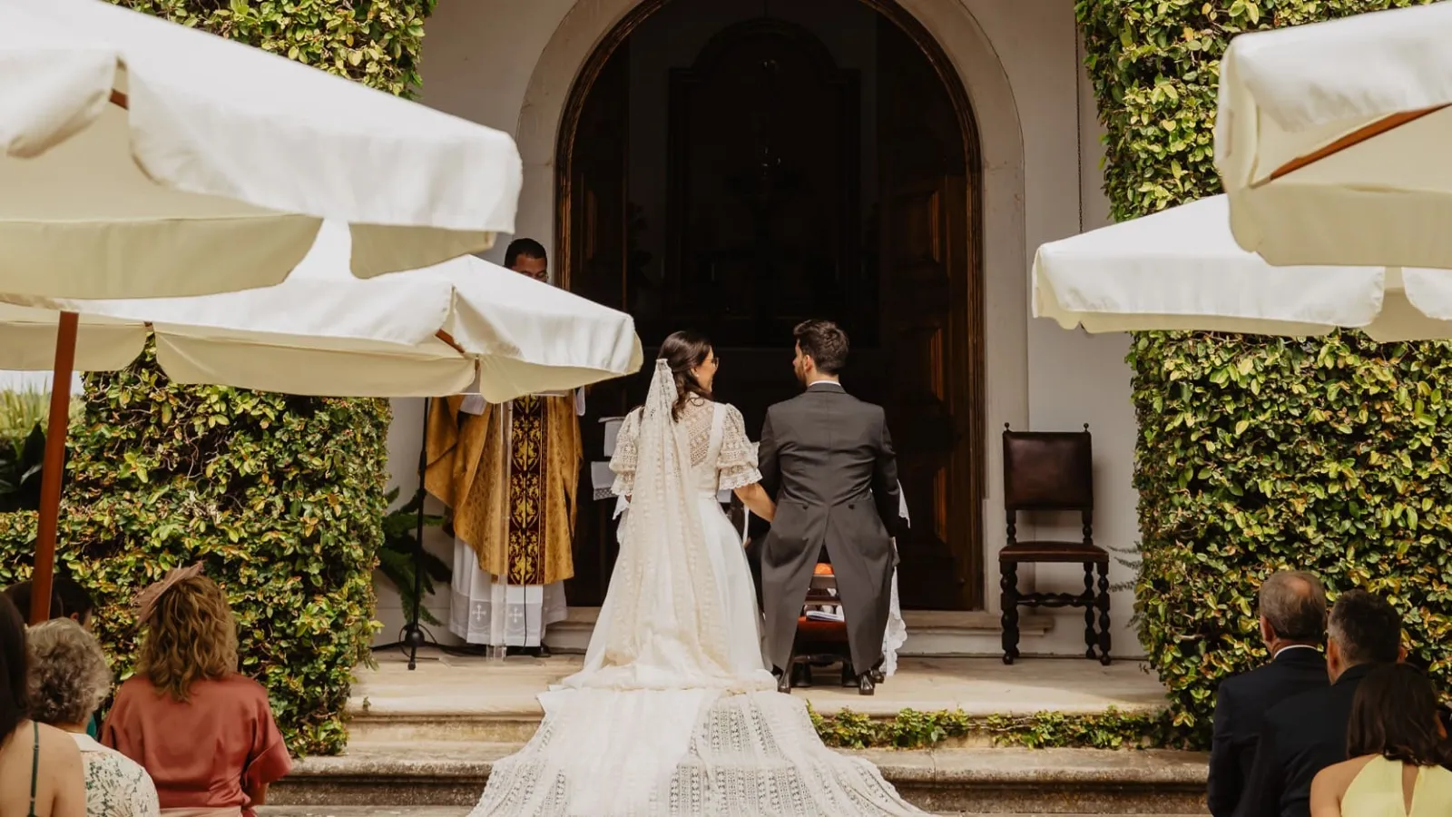 Cerimónia de casamento ao ar livre, com os noivos de costas em frente à capela